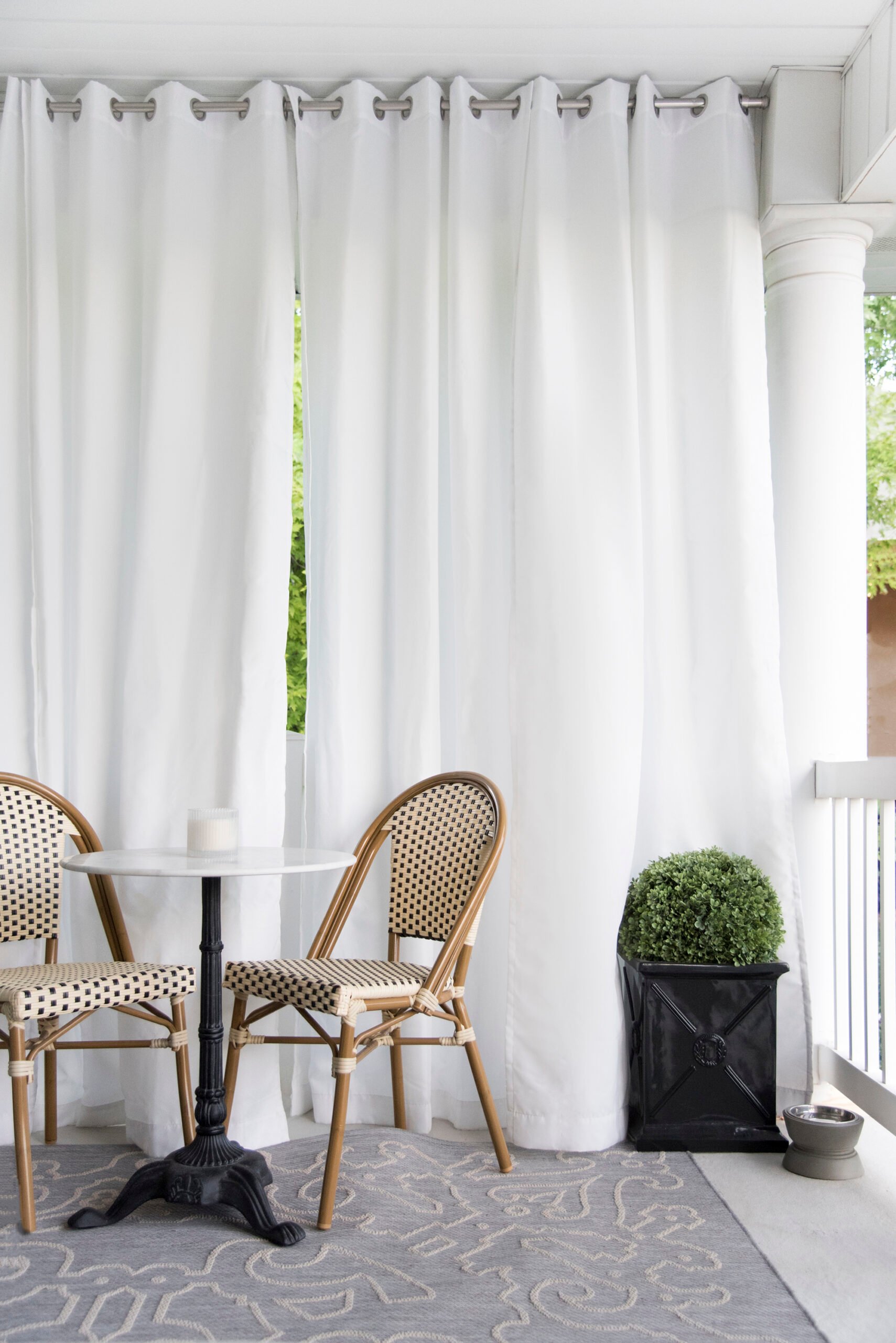 Balcony bistro table with woven chairs, a boxwood planter, and outdoor curtains