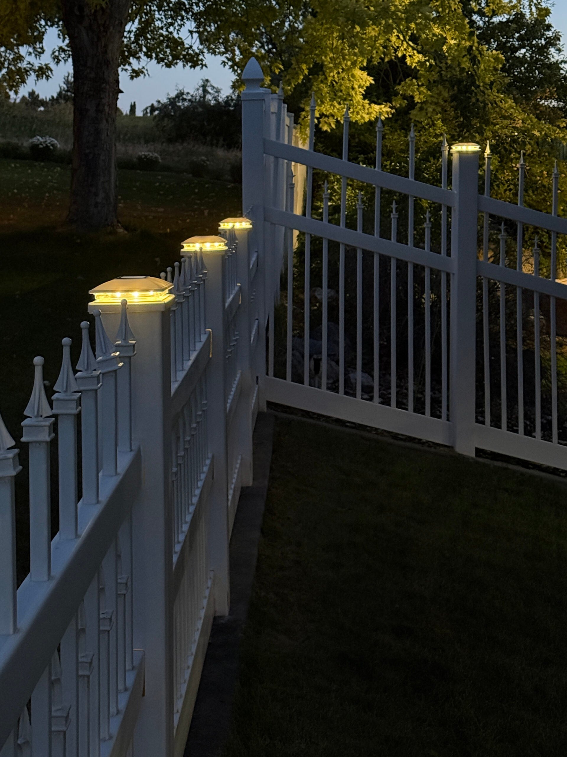 Illuminated white vinyl fence post row at night with solar post cap lights with a warm glow in a summer backyard