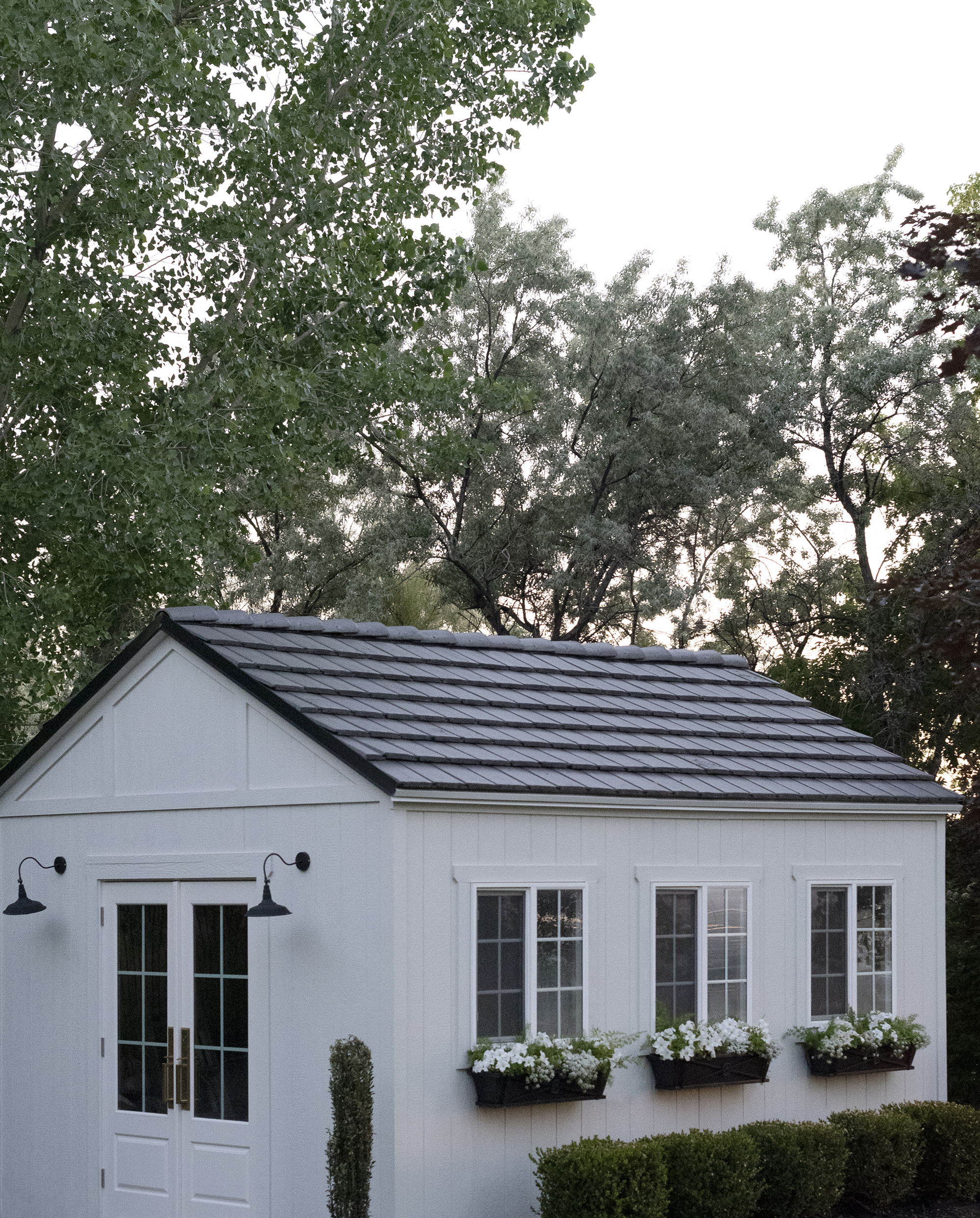 White storage shed with tile shingles, flower box planters with white blooms, and a row of boxwoods at the corner of a yard at dusk.