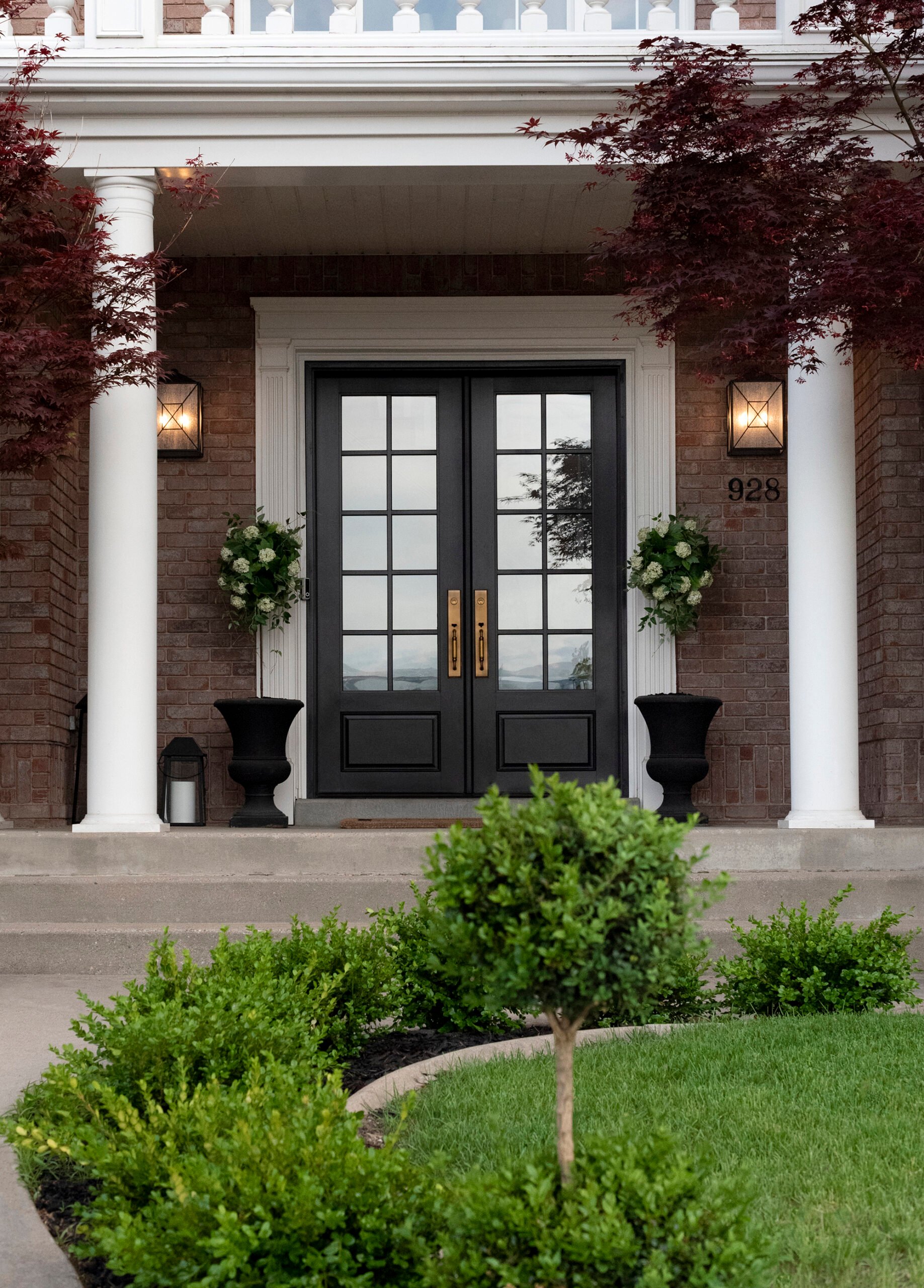 Exterior of Colonial home with boxwoods lining the sidewalk path, black french doors with brass hardware, and a classic brick facade. The doors are flanked by two topiary planters.