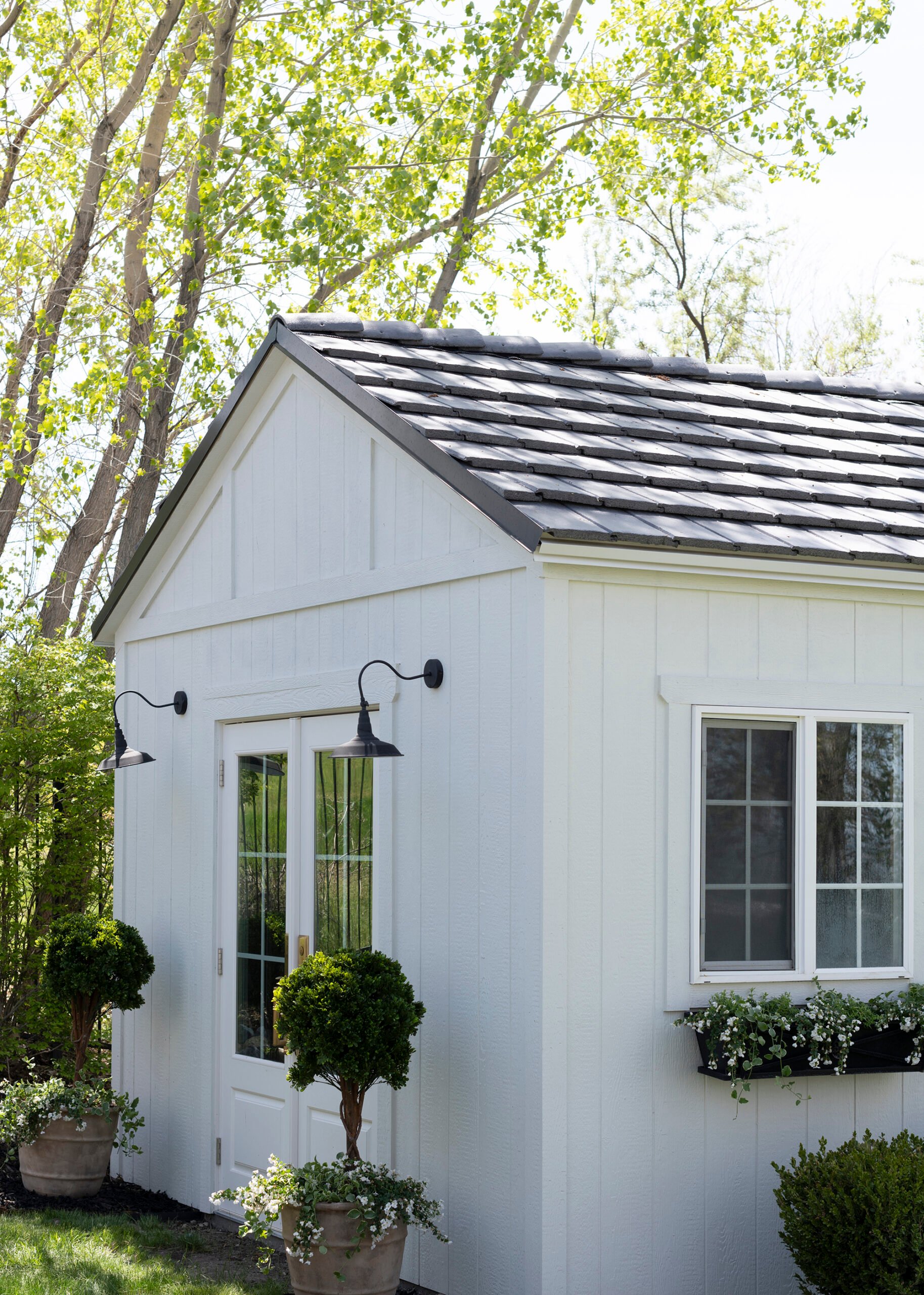 White storage shed with slate roof, window planter boxes, and topiaries flanking white french doors